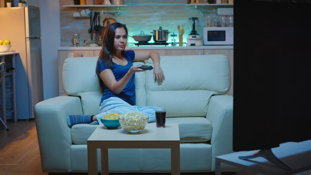 Woman seated on a couch, holding a TV remote, with snacks on the table in front of her