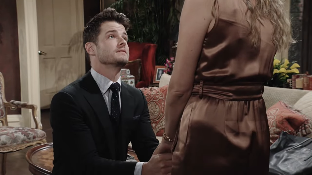 Kyle Abbott in classic suit standing on his kneel in front of women in silk brown dress